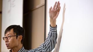 A side profile image of an asian man standing in front of a whileboard teaching a class.